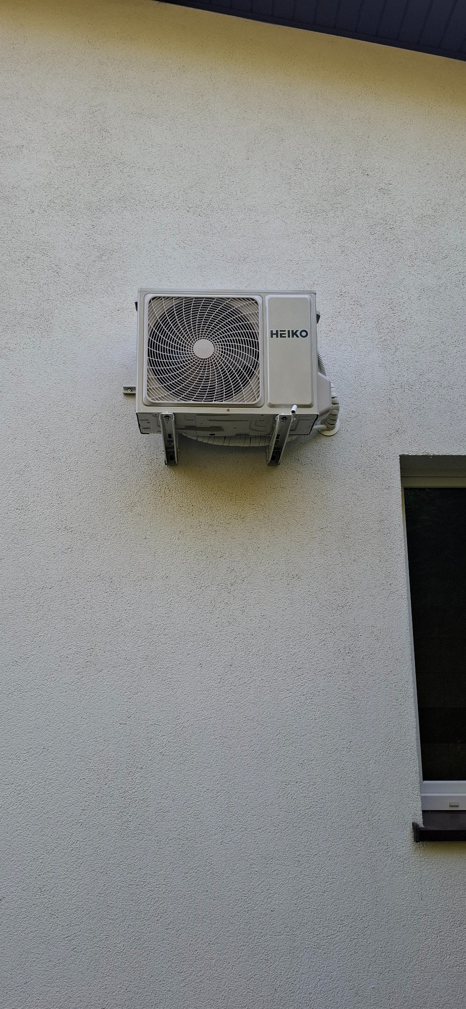 Zamontowana jednostka zewnętrzna klimatyzacji HEIKO na jasnej, teksturowanej ścianie budynku, widoczna rama okienna w prawym dolnym rogu.