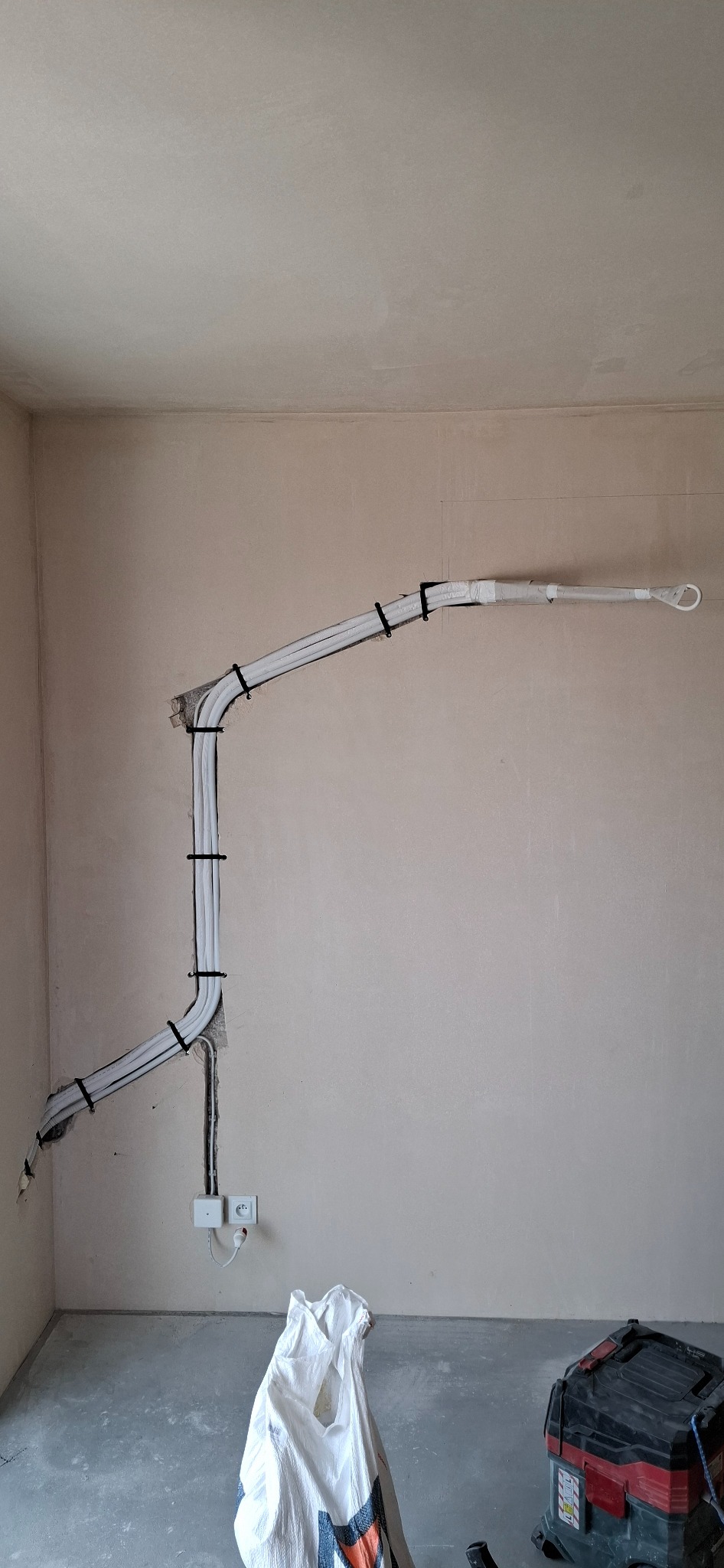 Białe rury instalacji, przymocowane czarnymi opaskami do ściany, połączone z gniazdkiem elektrycznym. Na podłodze worek i odkurzacz.