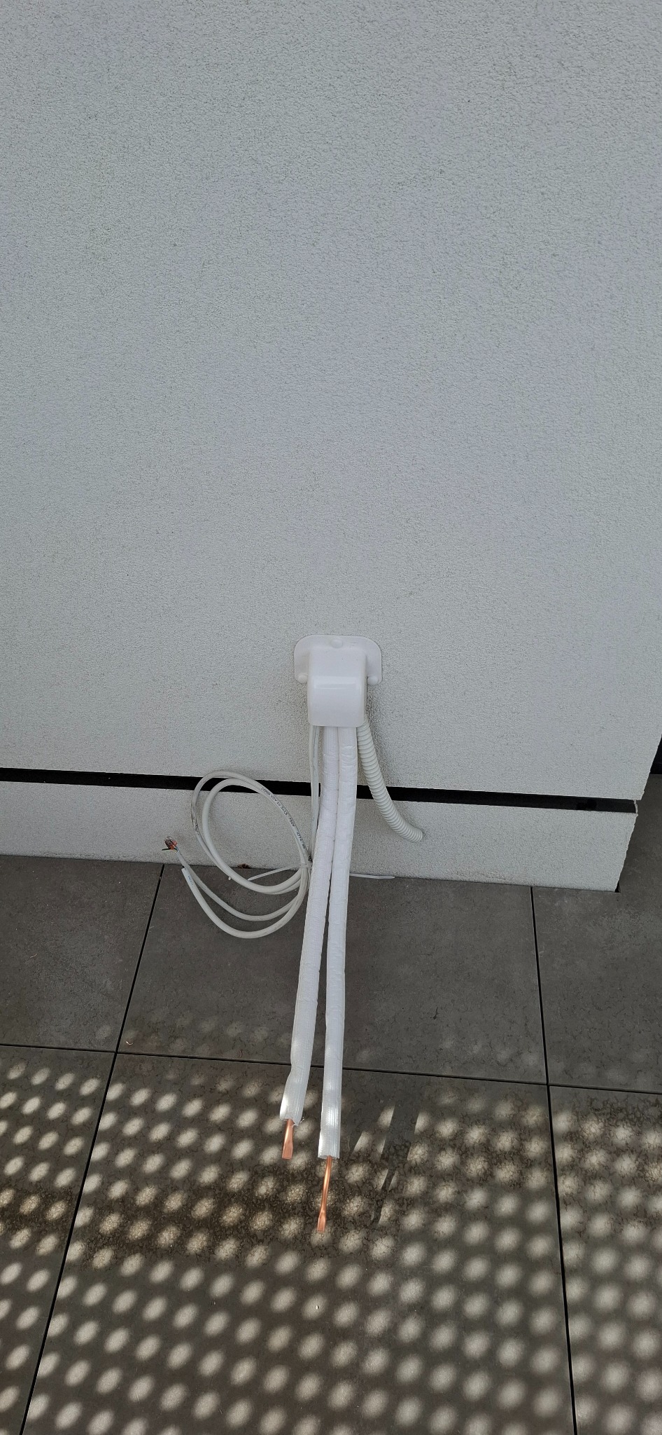 Biała instalacja klimatyzacji wychodząca ze ściany, z widocznymi miedzianymi rurkami i przewodami elektrycznymi na tle szarej podłogi i białej ściany.