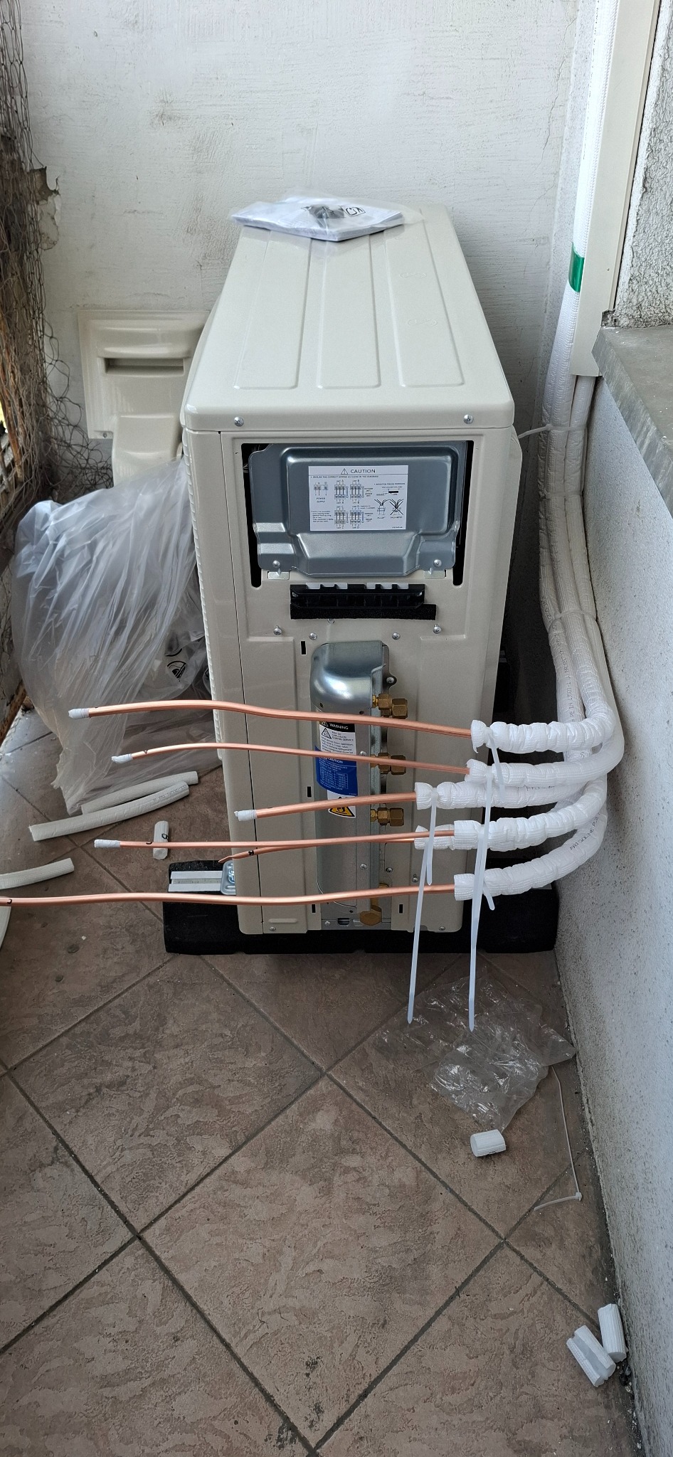 Widok z tyłu nowo zainstalowanej jednostki zewnętrznej klimatyzacji z podłączonymi rurami miedzianymi, częściowo zaizolowanymi, na kafelkowanym balkonie.