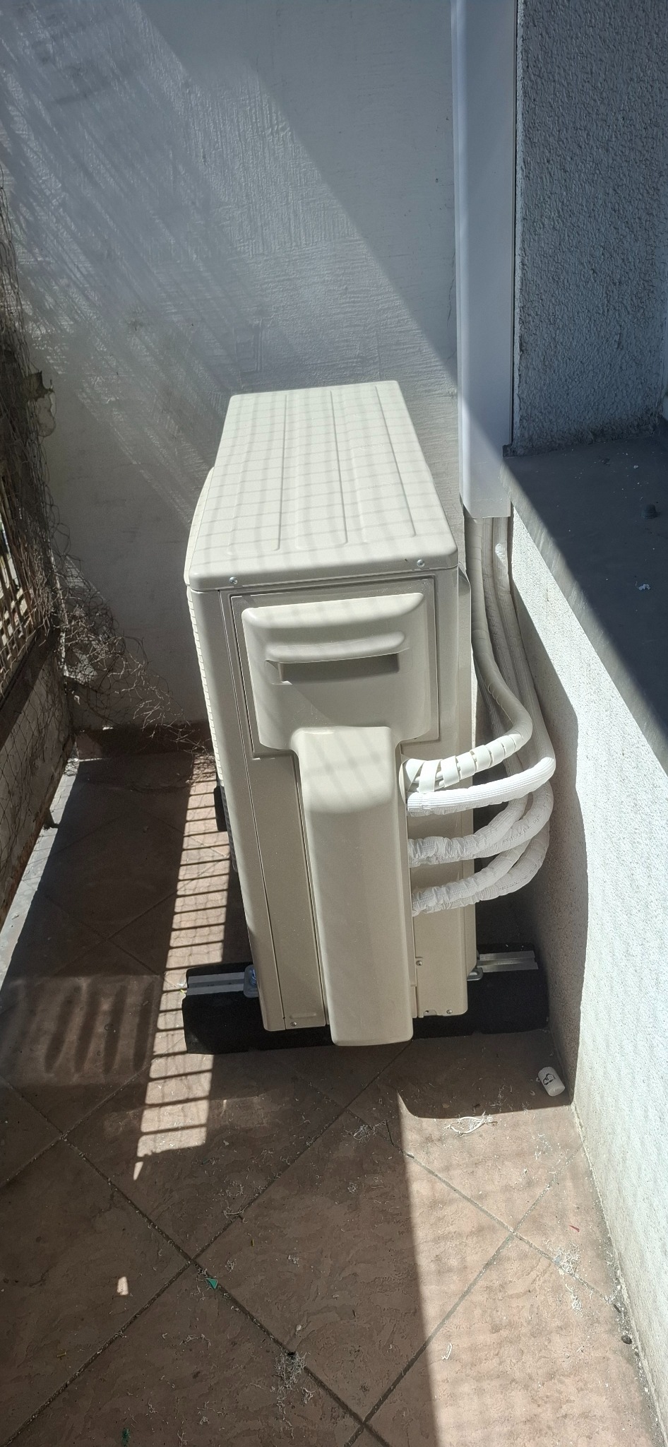 Zewnętrzna jednostka klimatyzacji zamontowana na balkonie, z widocznymi rurami w osłonie. Cień rzuca krata balkonu. Ujęcie z góry.