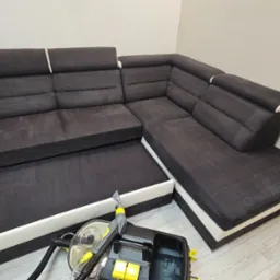 Narożnik z ciemną tapicerką i jasnymi wstawkami poddawany czyszczeniu ekstrakcyjnemu przy użyciu profesjonalnego odkurzacza piorącego z żółtymi elementami.