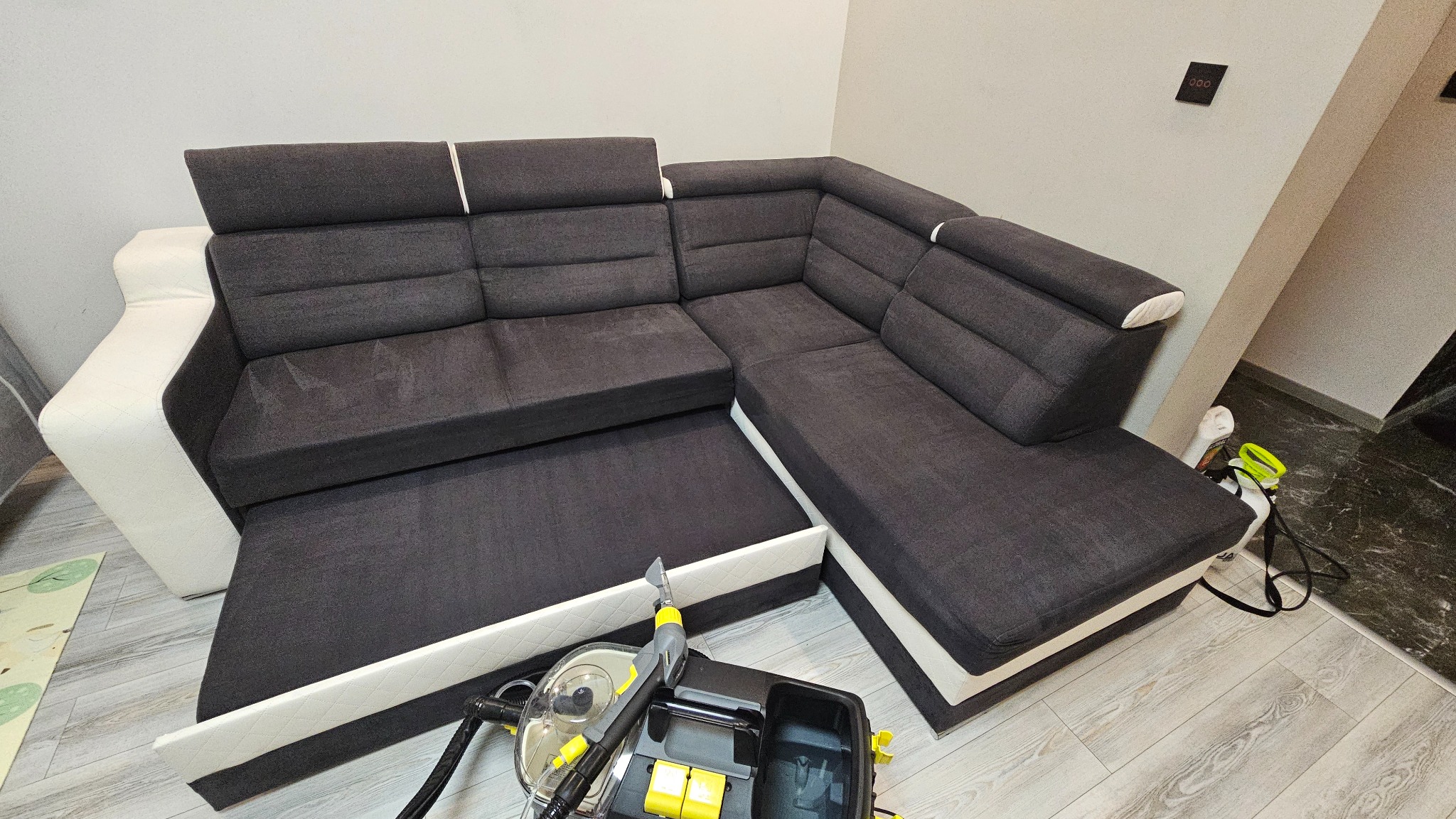 Narożnik z ciemną tapicerką i jasnymi wstawkami poddawany czyszczeniu ekstrakcyjnemu przy użyciu profesjonalnego odkurzacza piorącego z żółtymi elementami.