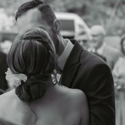 Sandra Marcinkowska - Czarno-białe zdjęcie obejmującej się pary, z widocznym tyłem głowy mężczyzny z brodą i upięciem włosów kobiety z kwiatem, w tle rozmyte sylwetki gości i samochód.