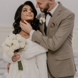 Sandra Marcinkowska - Narzeczona w białej sukni z bukietem białych róż, narzeczony w beżowym garniturze z muszką, delikatne ujęcie pary w jasnym wnętrzu.