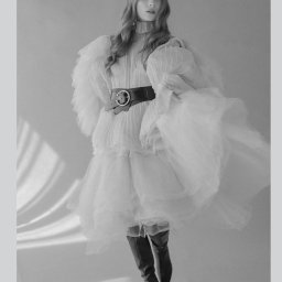 Sandra Marcinkowska - Czarno-białe zdjęcie modelki w plisowanej, białej sukience z bufiastymi rękawami, podkreślonej szerokim, skórzanym paskiem z okrągłą klamrą i wysokimi, skórzanymi kozakami, na jasnoszarym tle...