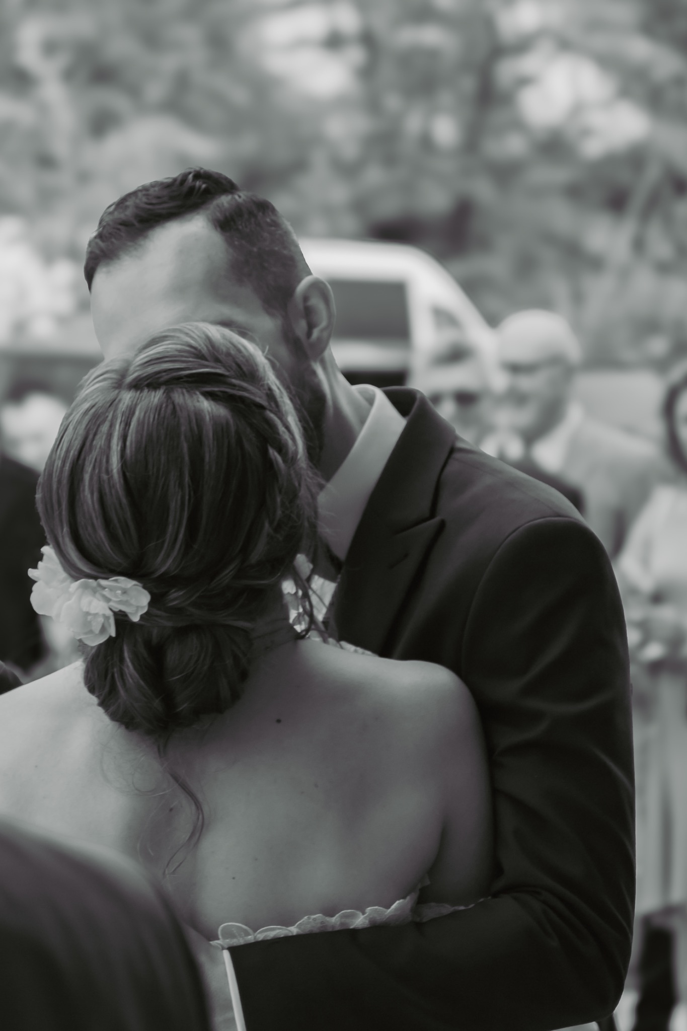 Czarno-białe zdjęcie obejmującej się pary, z widocznym tyłem głowy mężczyzny z brodą i upięciem włosów kobiety z kwiatem, w tle rozmyte sylwetki gości i samochód.