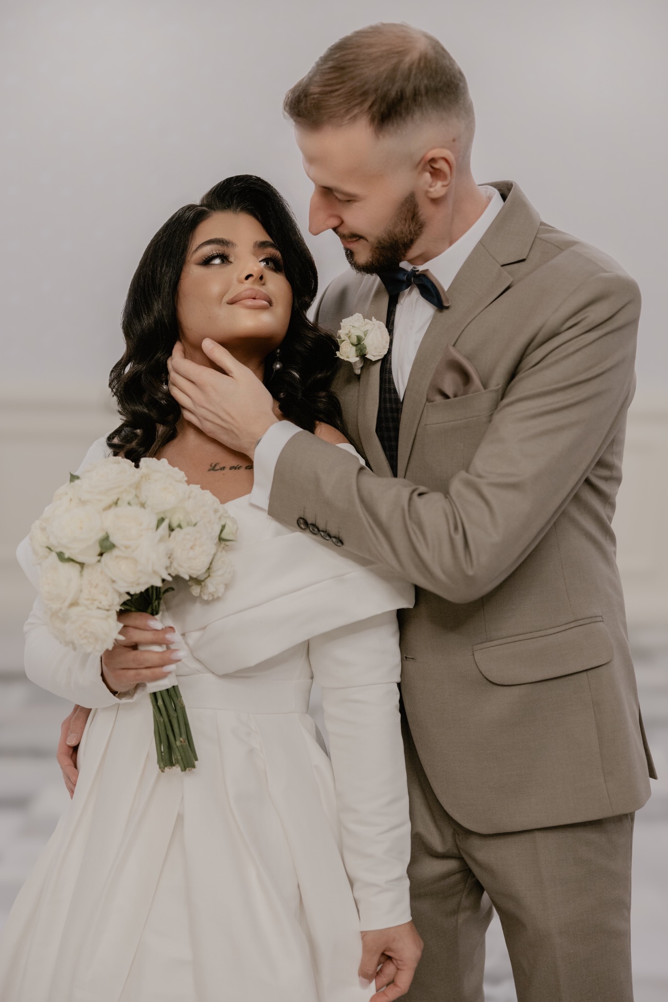 Narzeczona w białej sukni z bukietem białych róż, narzeczony w beżowym garniturze z muszką, delikatne ujęcie pary w jasnym wnętrzu.