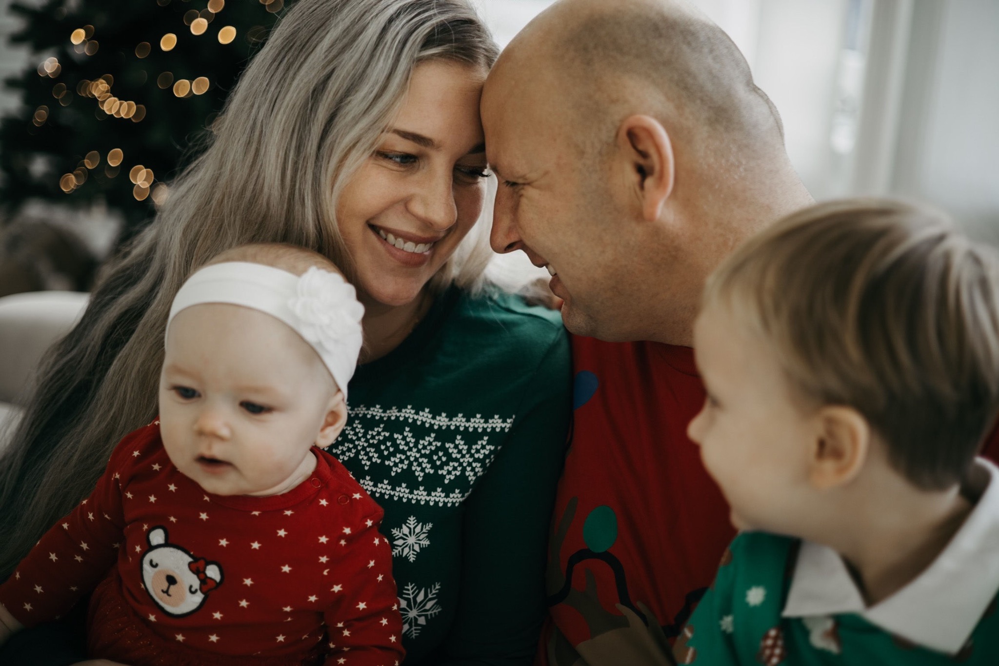 Rodzinne zbliżenie: mama z blond włosami i tata dotykają się czołami, uśmiechają się, obok nich dwoje małych dzieci w świątecznych ubraniach, w tle choinka z delikatnym bokeh.