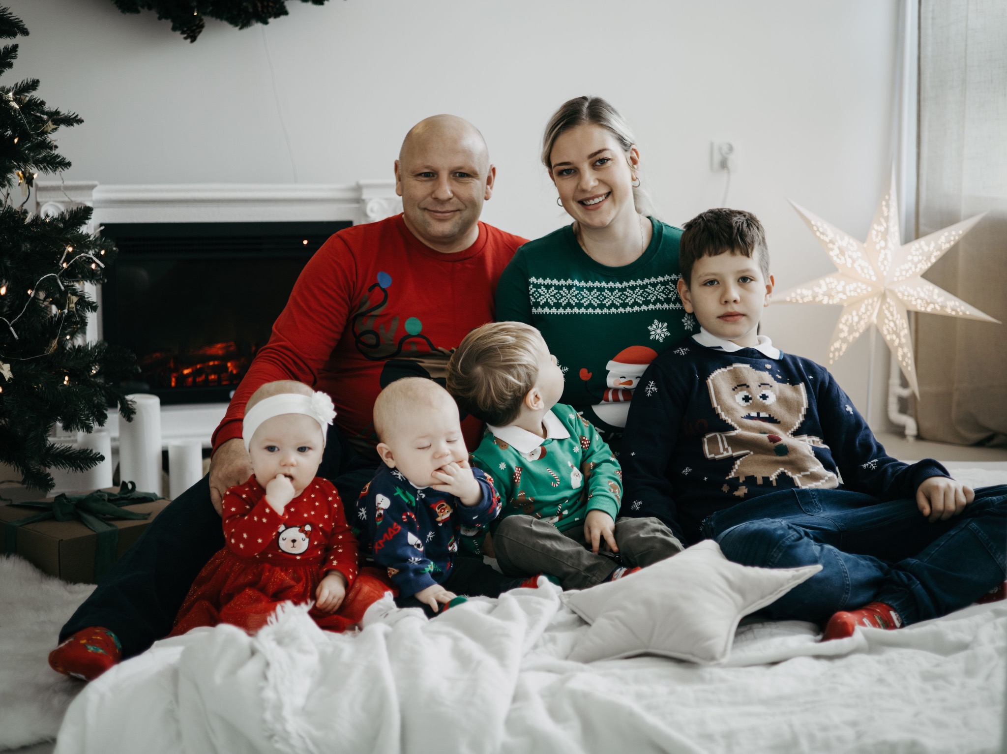 Rodzinne zdjęcie w świątecznych swetrach, z trzema małymi dziećmi i choinką w tle, na białym kocu z poduszkami w kształcie gwiazd.