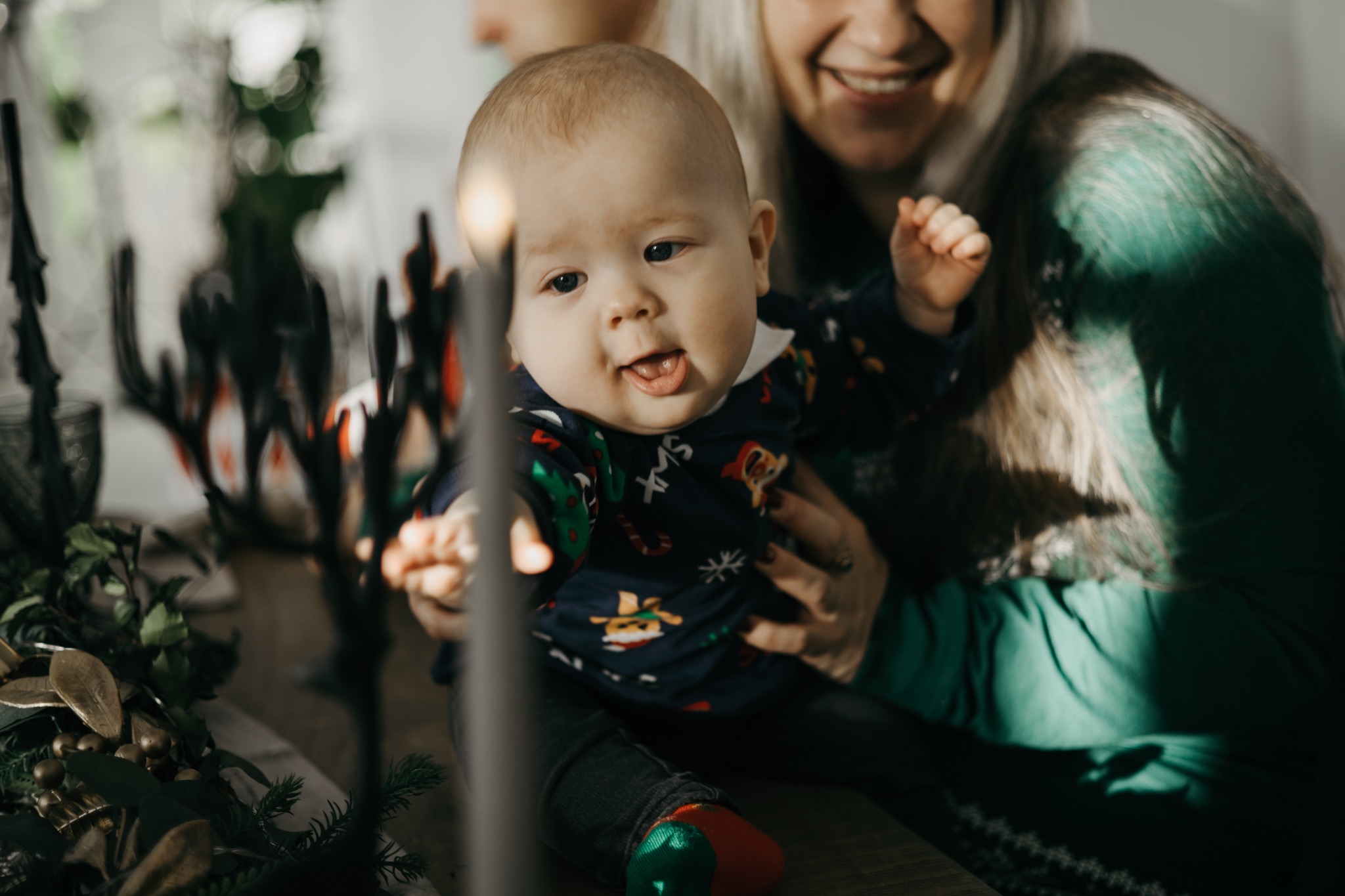 Uśmiechnięty niemowlak w świątecznym ubranku na kolanach kobiety, z dekoracyjną świecą i zielonymi gałązkami w tle.