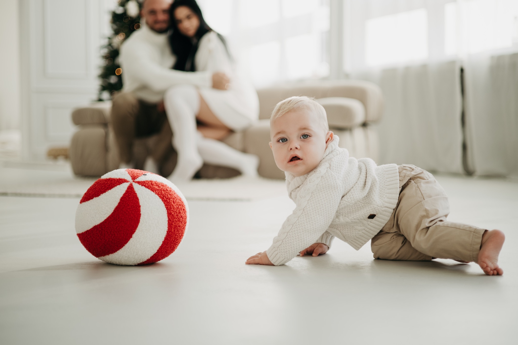 Pełzający chłopiec w beżowych spodniach i białym swetrze, z czerwoną piłką w paski, w tle para siedząca na kanapie przy choince w Opolu.