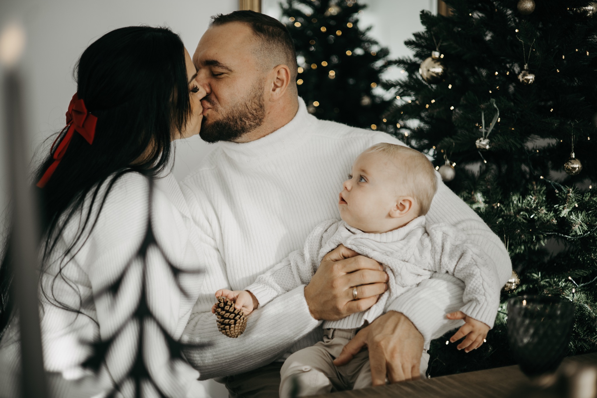 Rodzinne zdjęcie w świątecznej atmosferze: całująca się para w białych swetrach, trzymająca na rękach małe dziecko z szyszką w dłoni, choinka w tle.