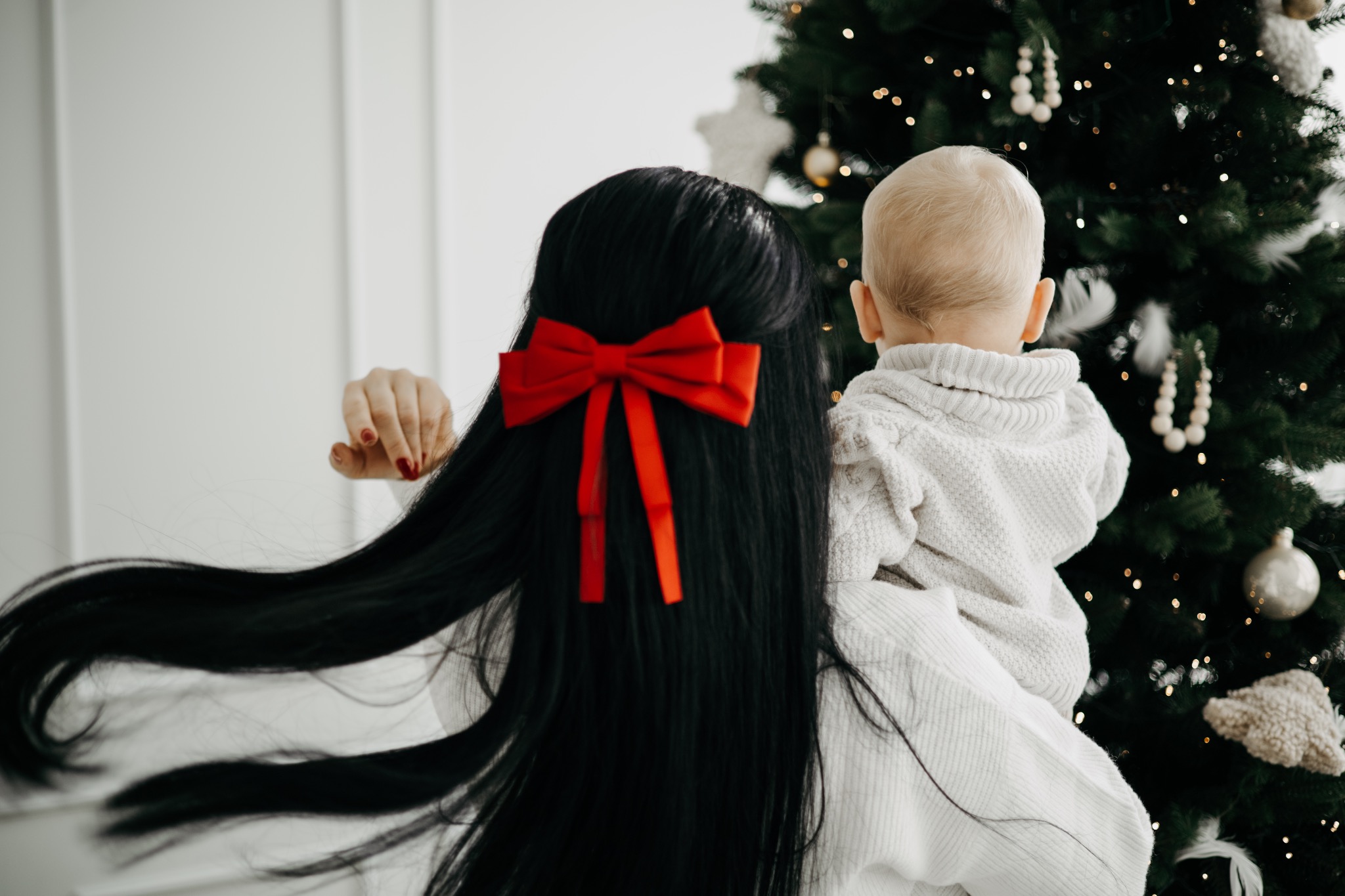 Kobieta z długimi czarnymi włosami i czerwoną kokardą trzyma na rękach małe dziecko, oboje stoją tyłem do aparatu na tle choinki z białymi ozdobami i delikatnymi światełkami.