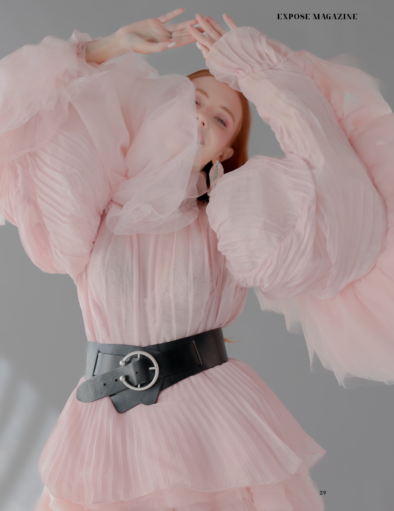 Kobieta w plisowanej, różowej sukience z bufkami i szerokim czarnym pasem, pozująca z rękami uniesionymi nad głową, widoczna częściowo twarz i kolczyki.