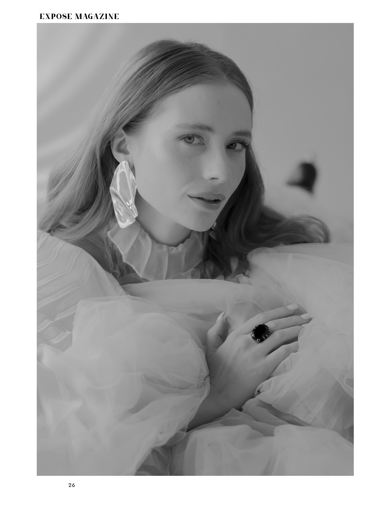 Portret kobiety w czarno-białej tonacji, z dużym asymetrycznym kolczykiem i pierścieniem z czarnym kamieniem, otulona przezroczystą tkaniną z falbanami.