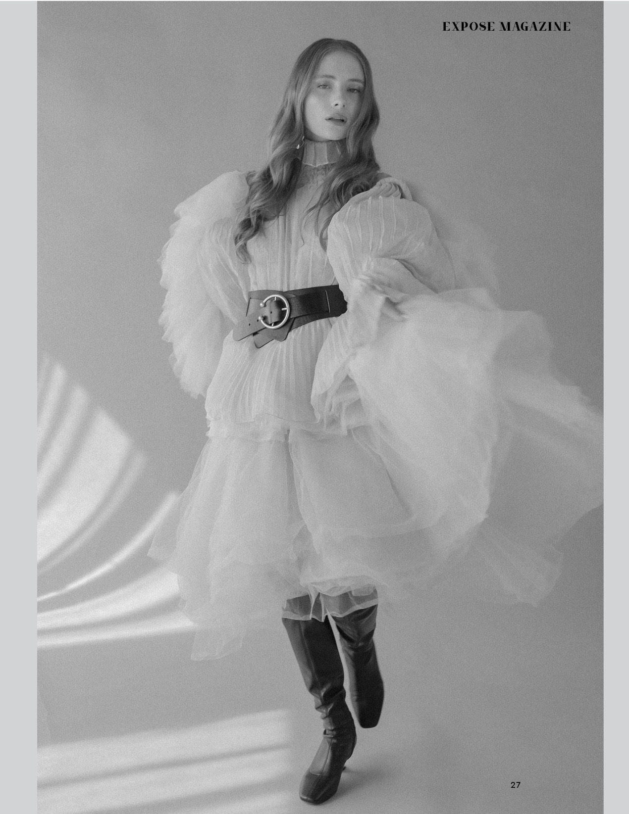 Czarno-białe zdjęcie modelki w plisowanej, białej sukience z bufiastymi rękawami, podkreślonej szerokim, skórzanym paskiem z okrągłą klamrą i wysokimi, skórzanymi kozakami, na jasnoszarym tle...