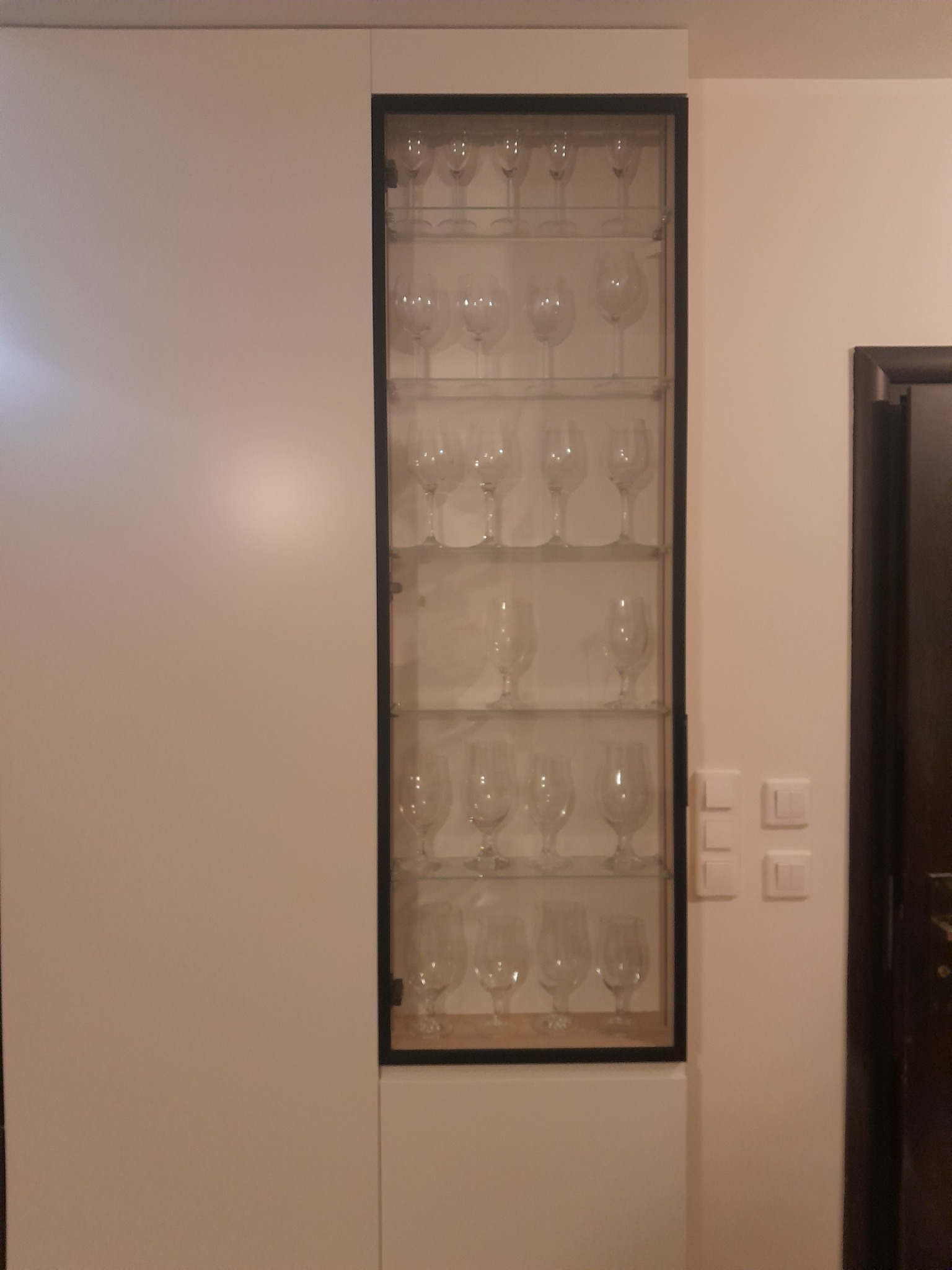 Biała szafa na wymiar z przeszkloną witryną, prezentująca kolekcję kieliszków w Elblągu. Minimalistyczny design, proste linie, czarna rama witryny.