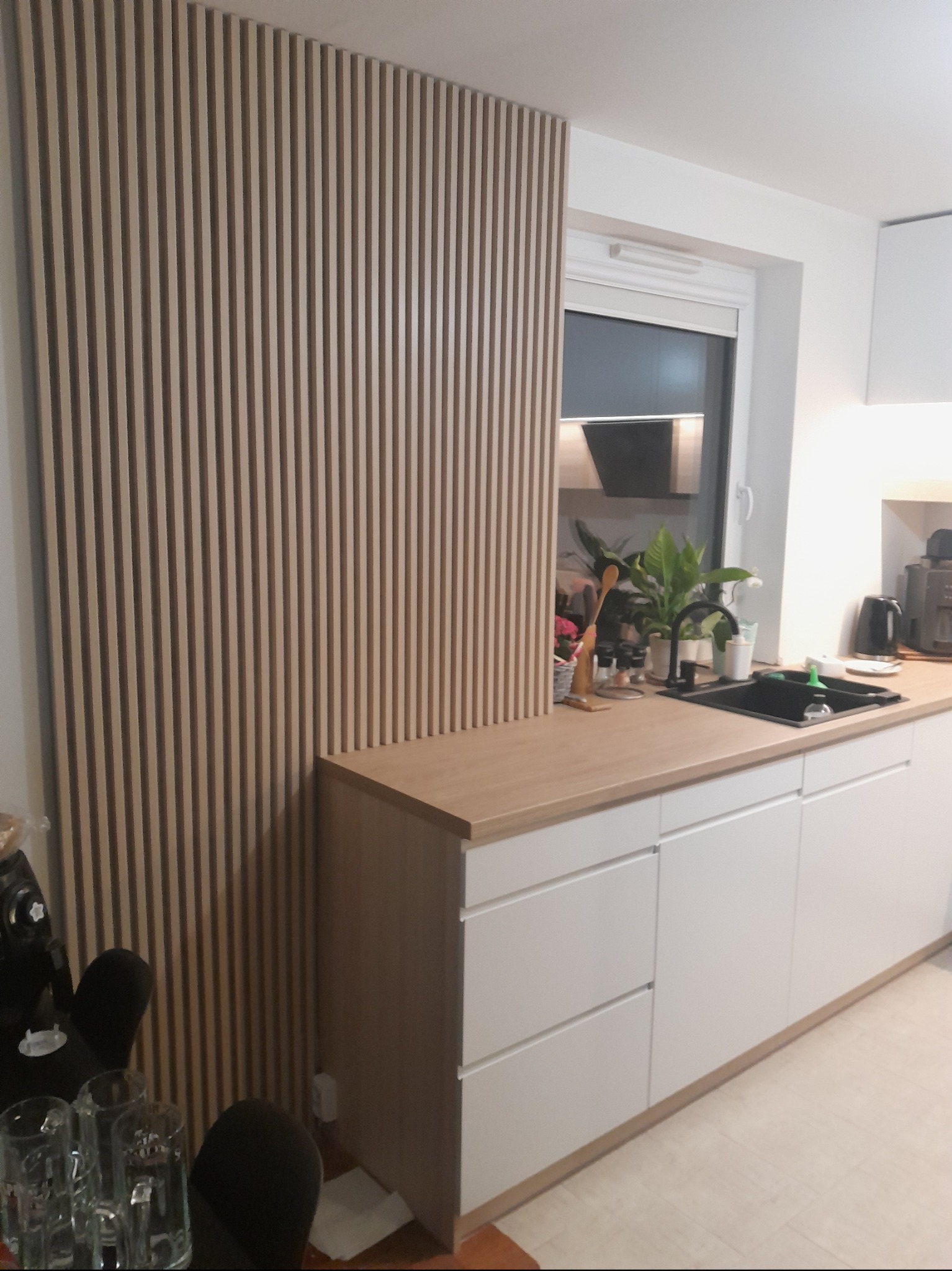 Nowoczesna kuchnia z białymi szafkami i blatem w kolorze drewna, uzupełniona dekoracyjną ścianką z drewnianych listewek pionowych, zlew wpuszczany, widoczne rośliny doniczkowe na blacie przy oknie.