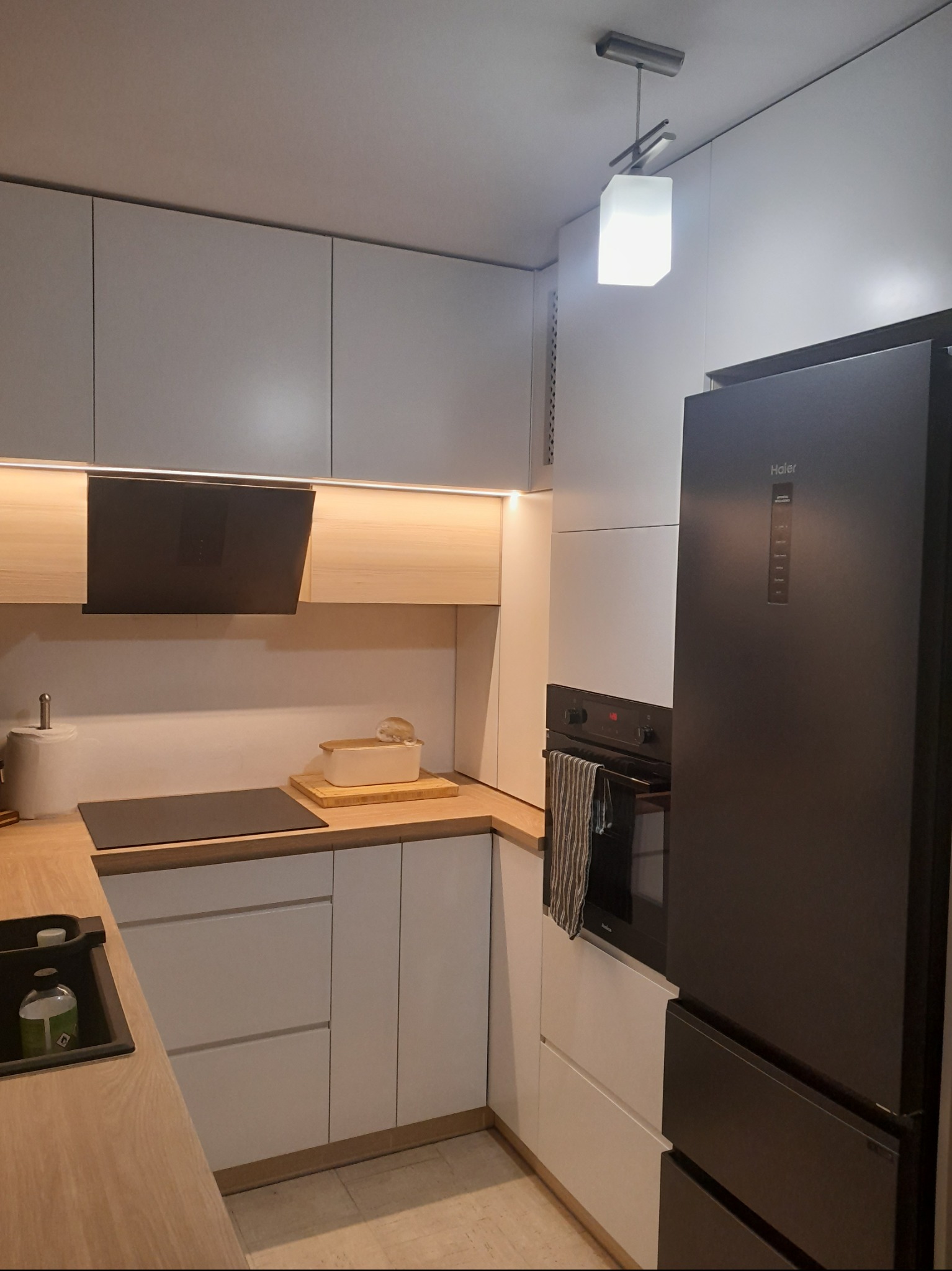 Nowoczesna, jasna kuchnia z szarymi górnymi szafkami, drewnianym blatem, czarną płytą indukcyjną i lodówką Haier.