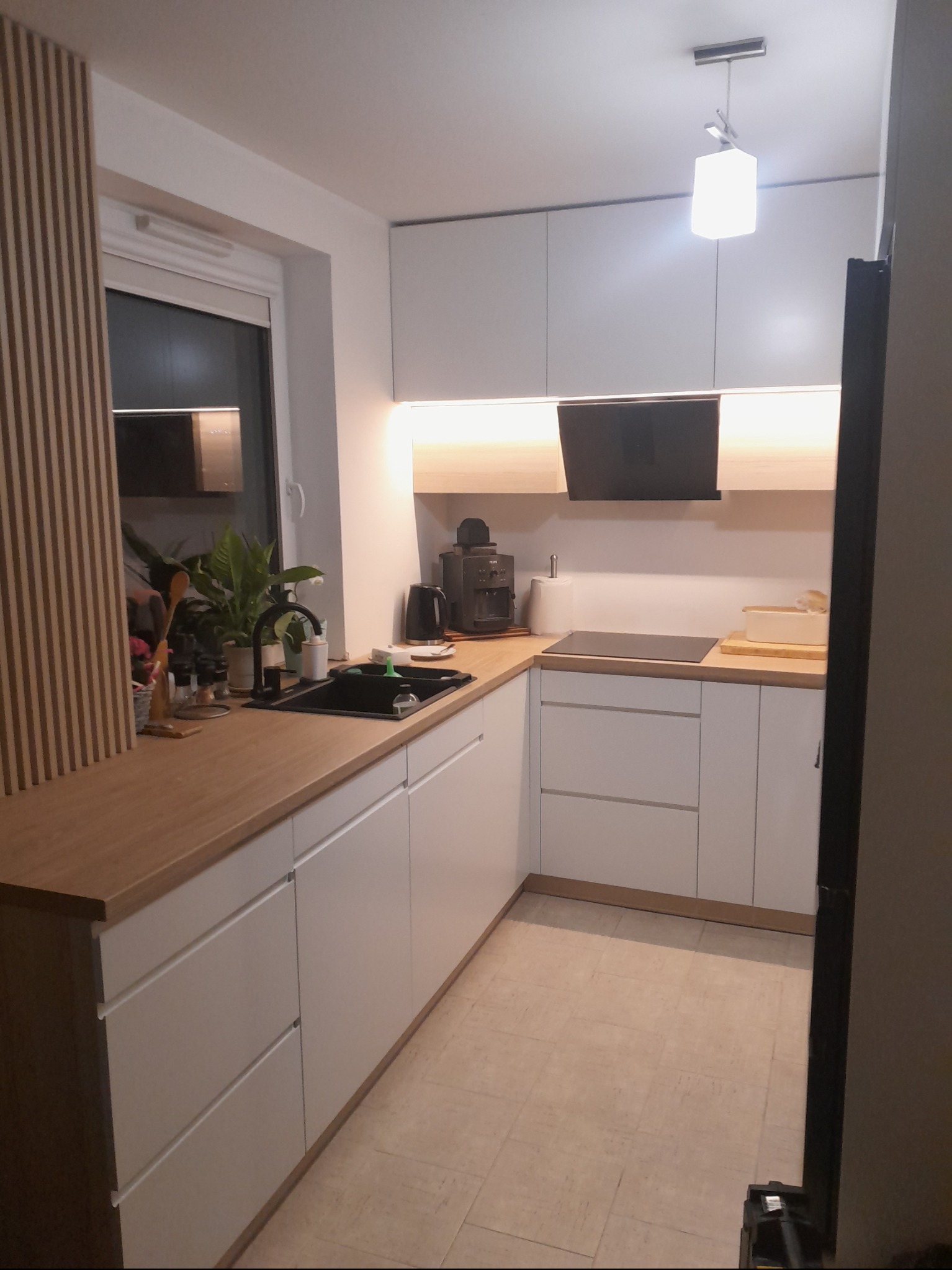 Nowoczesna kuchnia z białymi, matowymi frontami szafek, drewnianym blatem, czarnym zlewem i baterią, oraz dekoracyjną ścianką z drewnianych listewek przy oknie.