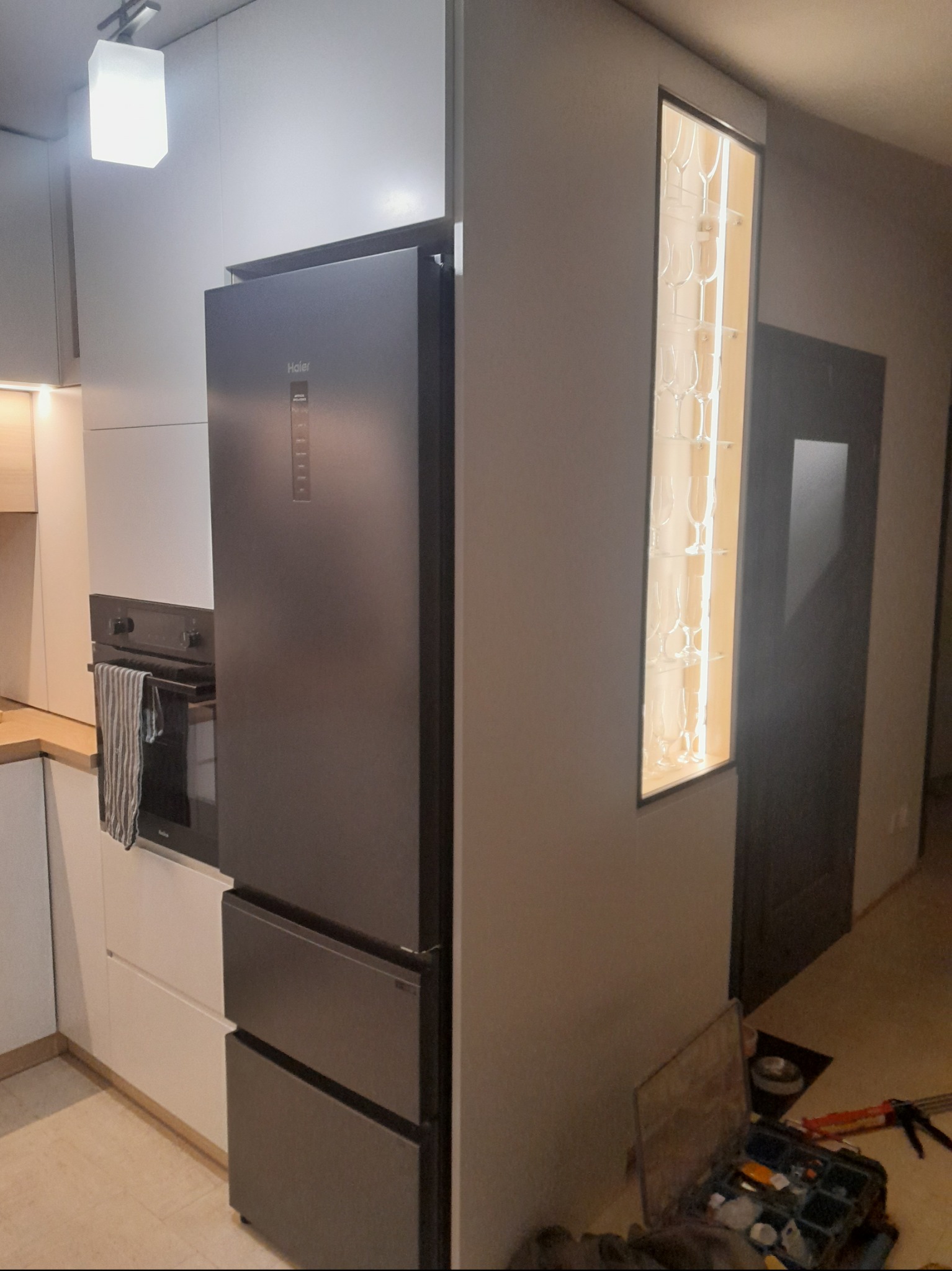 Nowoczesna kuchnia z zabudowaną lodówką Haier, oświetlona szafka na kieliszki i skrzynka narzędziowa na podłodze wskazują na trwający montaż.