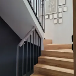 Nowoczesne schody drewniane z czarną balustradą, zabezpieczoną szarą osłoną, na ścianie galeria pustych ramek.