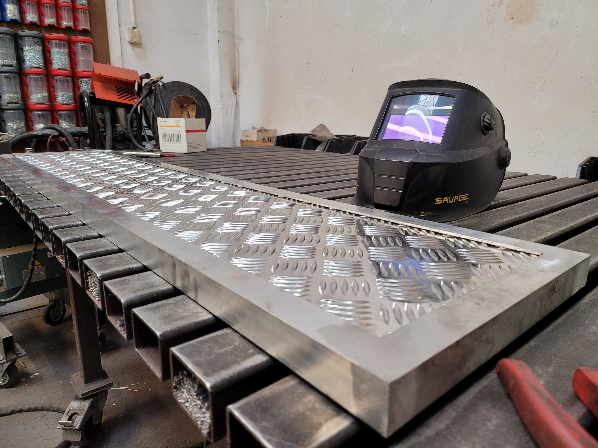 Aluminiowa płyta z wytłaczanym wzorem na metalowym stole warsztatowym obok maski spawalniczej Savage A-40