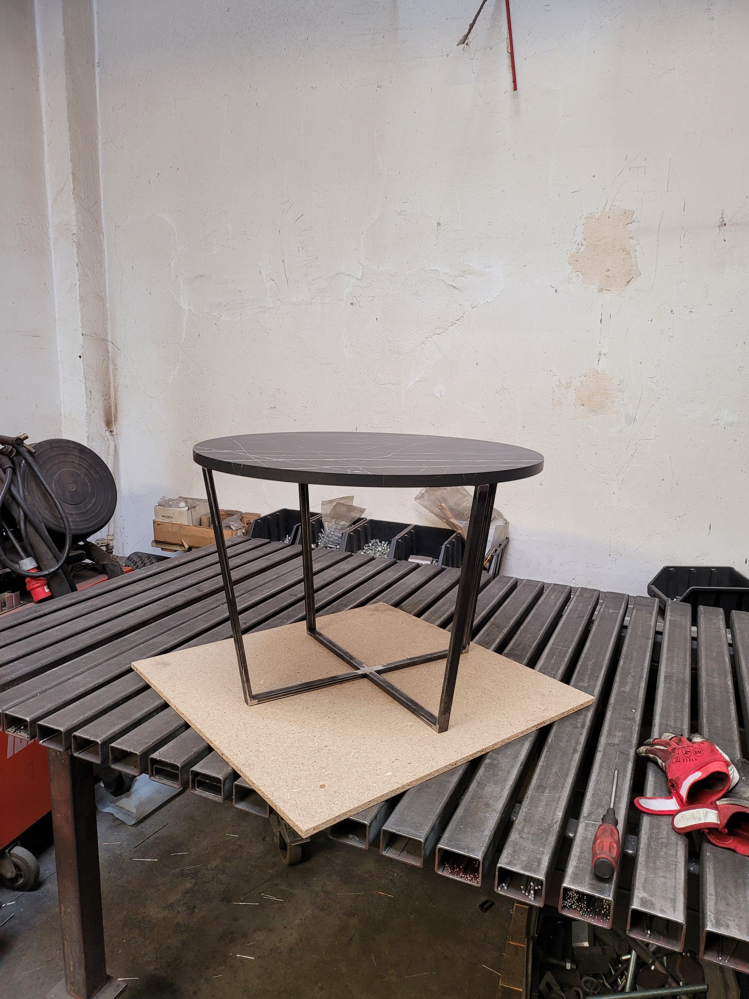 Stolik kawowy z okrągłym blatem imitującym marmur i metalową podstawą w trakcie montażu na stole spawalniczym, obok leżą rękawice robocze.