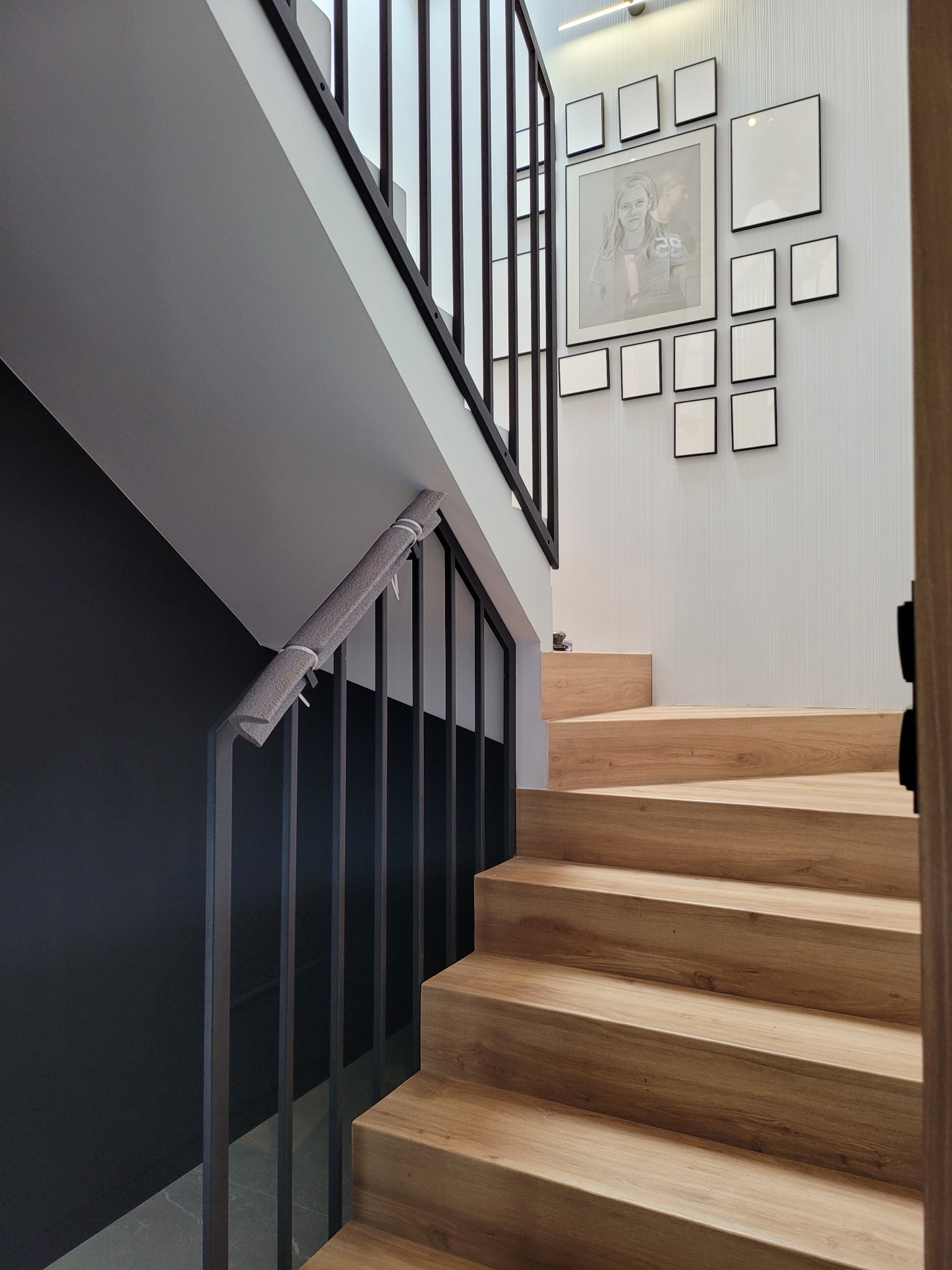 Nowoczesne schody drewniane z czarną balustradą, zabezpieczoną szarą osłoną, na ścianie galeria pustych ramek.