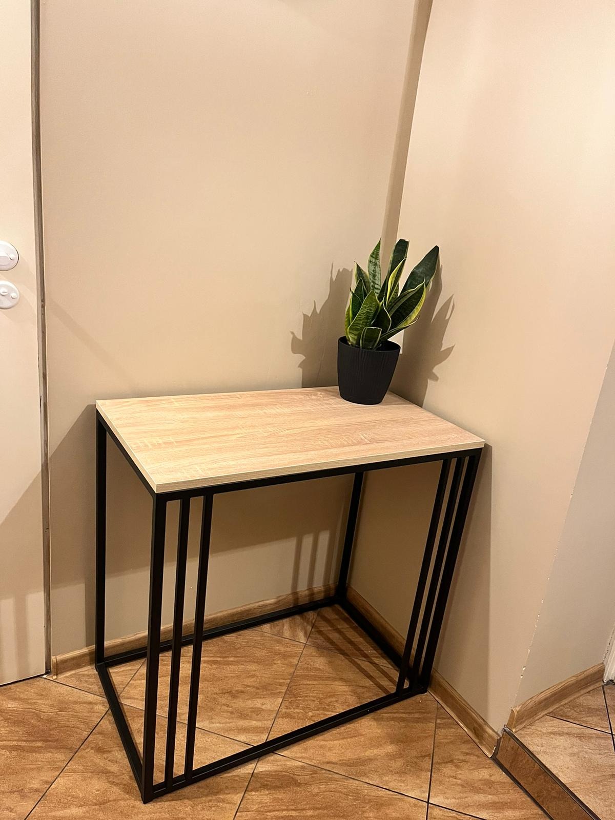 Stolik konsolowy z blatem imitującym jasne drewno i czarną, geometryczną podstawą z profili stalowych, na blacie doniczka z zieloną rośliną, ustawiony przy beżowej ścianie.