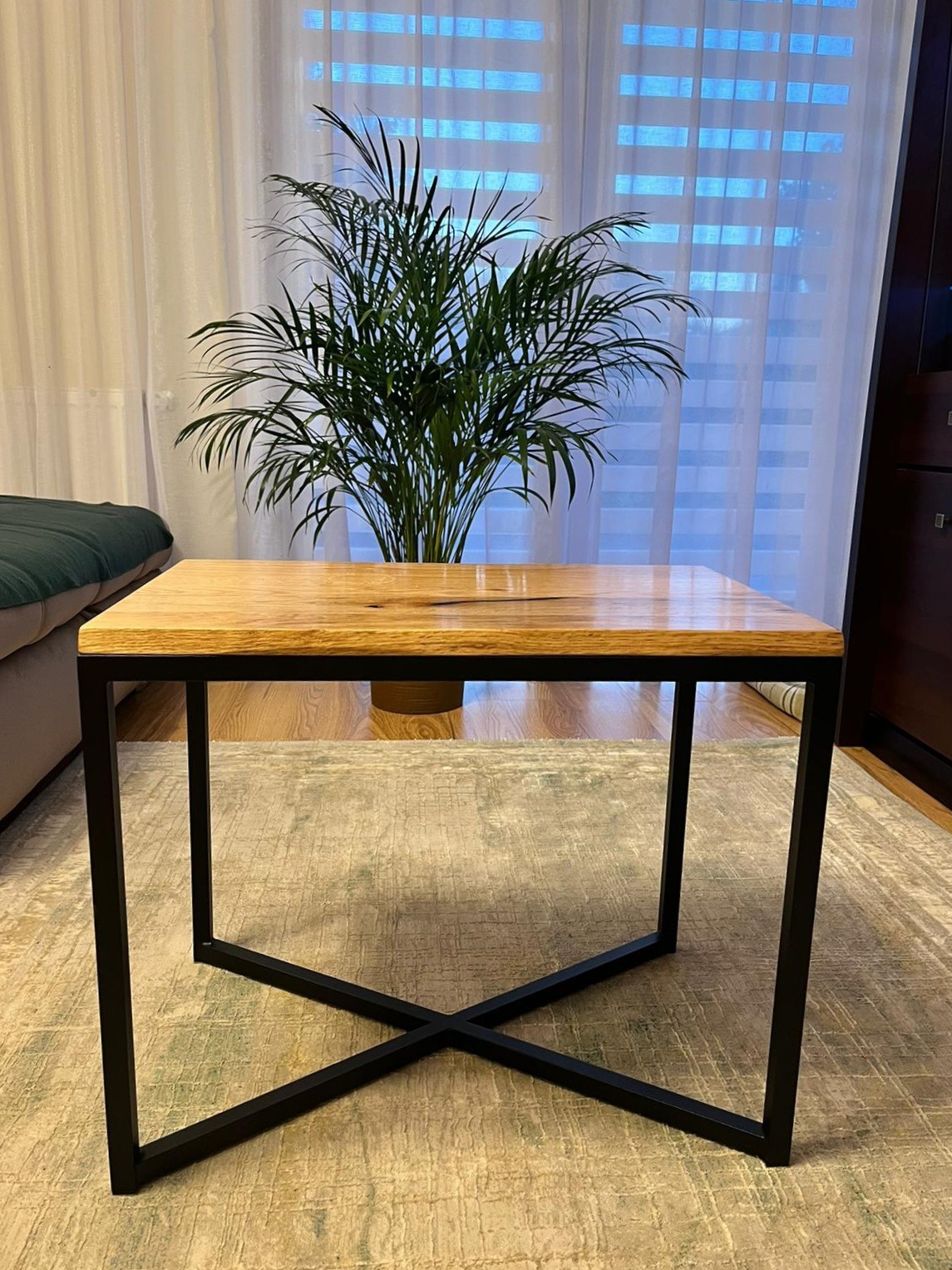 Stolik kawowy z blatem z jasnego drewna i czarną, metalową podstawą w kształcie litery X, na tle okna z roletami i rośliny doniczkowej.