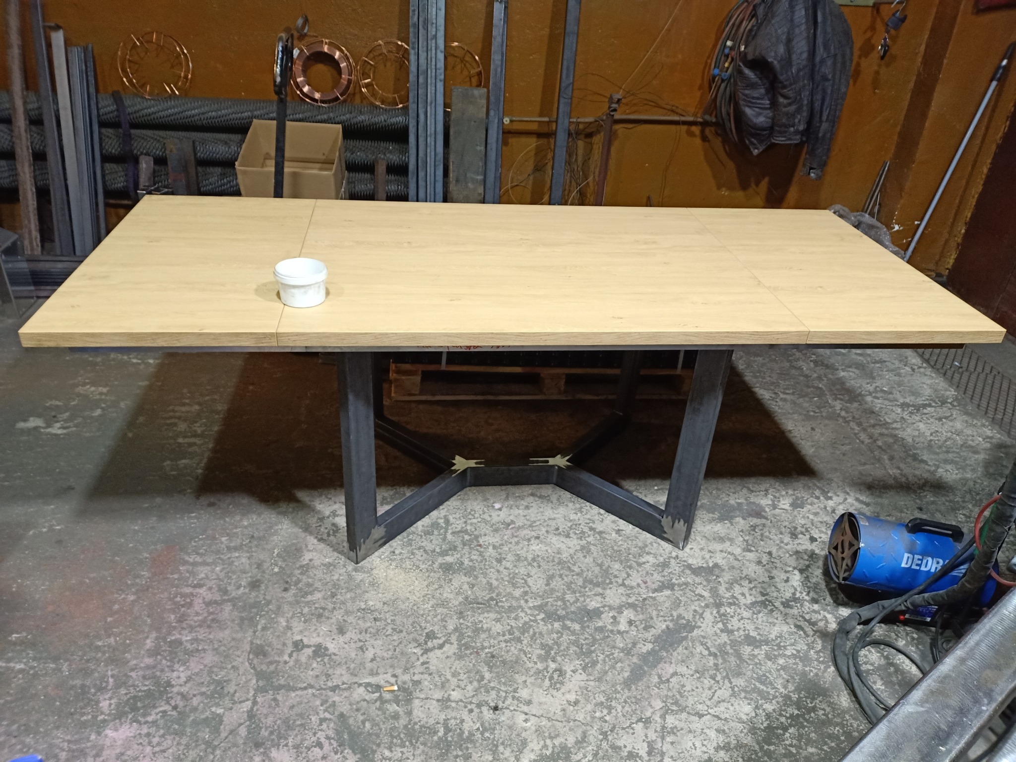 Stół z drewnianym blatem i spawaną, surową, metalową konstrukcją nóg w warsztacie, obok stoi nagrzewnica, w tle metalowe profile i zwinięte druty spawalnicze.