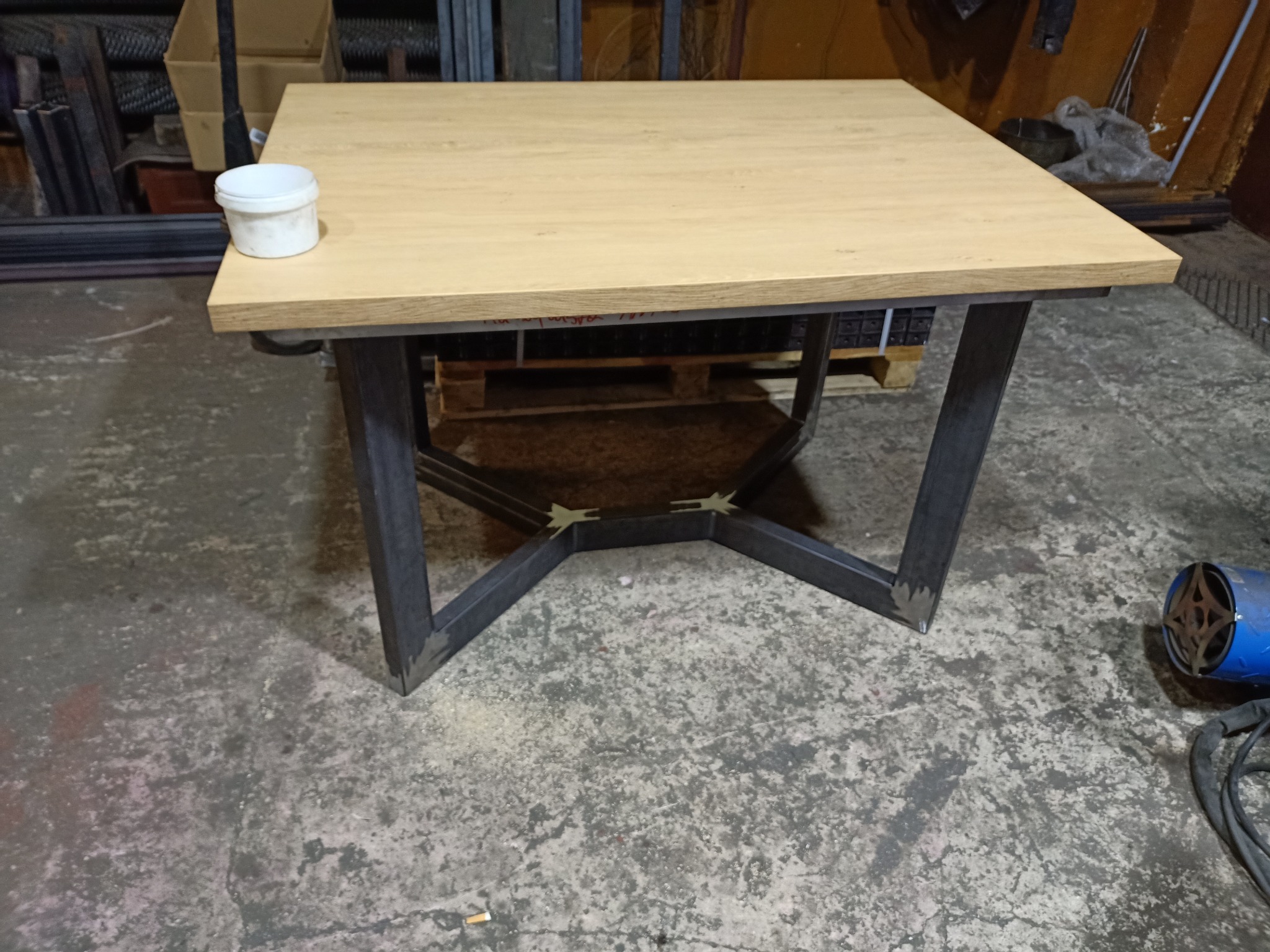 Stół z blatem imitującym jasne drewno i surową, spawaną, metalową podstawą w kształcie litery X, stojący na betonowej podłodze warsztatu, obok puszka z preparatem do drewna i spawarka.