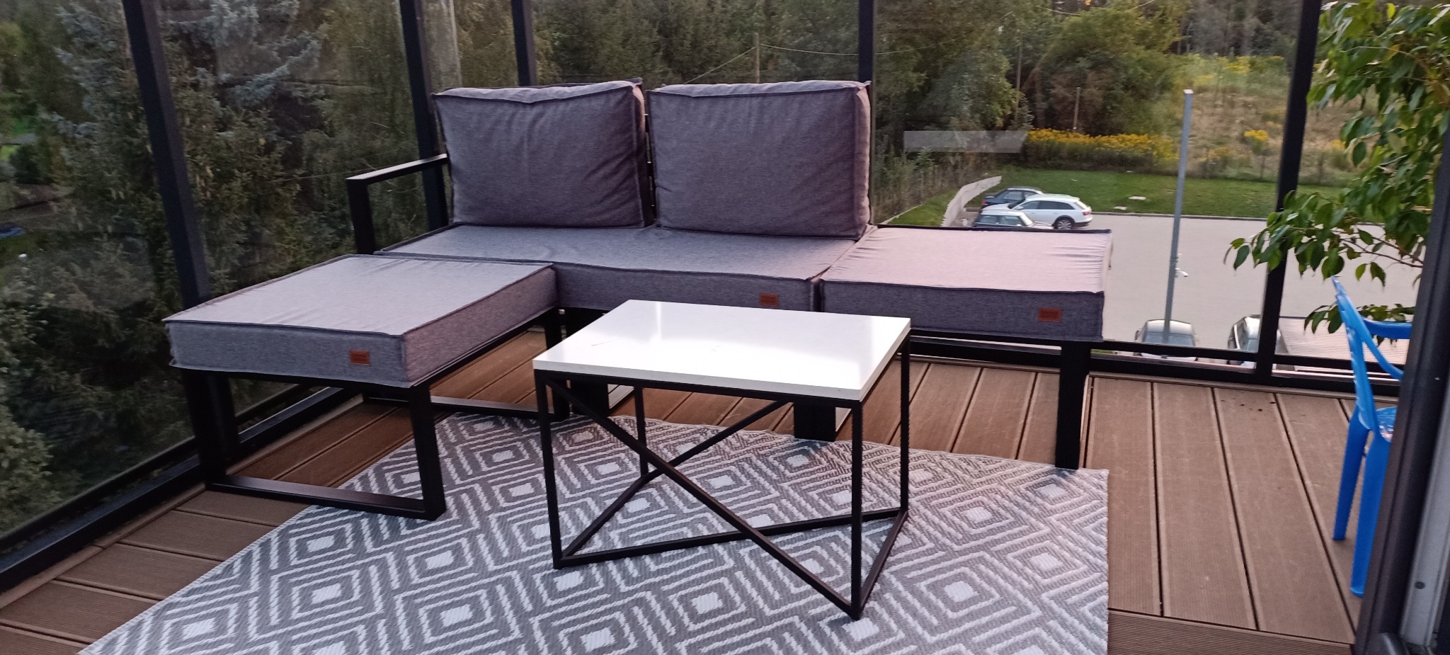 Zadaszony taras z narożną sofą o metalowej konstrukcji i szarych poduchach, stolikiem kawowym z białym blatem i wzorzystym dywanem.