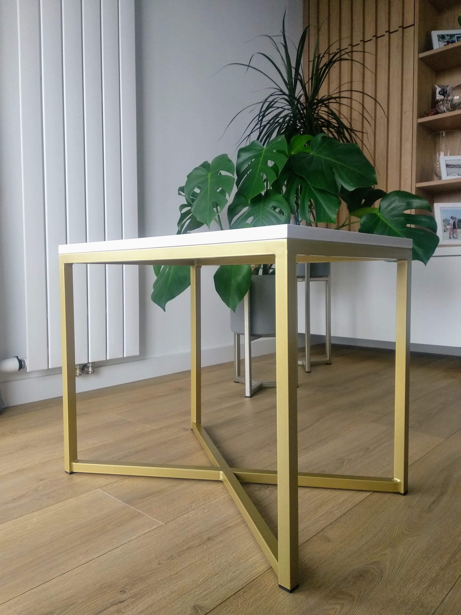 Stolik kawowy z białym blatem i złotymi, spawane ramami, na drewnianej podłodze, z roślinami w tle