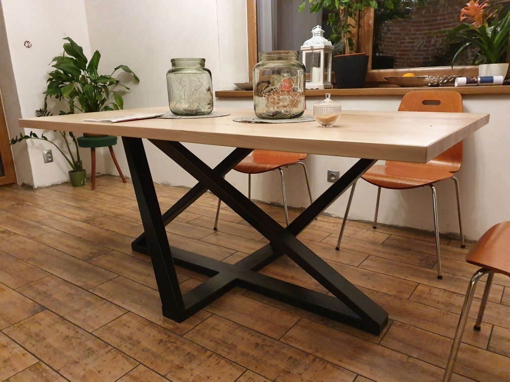 Stół z jasnym drewnianym blatem i czarną, geometryczną podstawą ze stali, otoczony krzesłami w ciepłych odcieniach brązu, ustawiony na podłodze z płytek imitujących drewno.