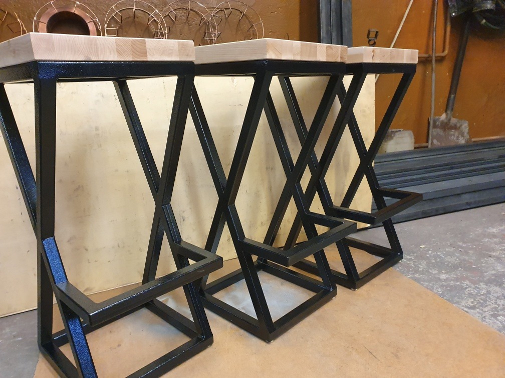 Trzy identyczne hokery z czarną metalową konstrukcją w kształcie litery X i jasnym, drewnianym siedziskiem, ustawione obok siebie na brązowym kartonie, w tle widoczne metalowe pręty i narzędzia.