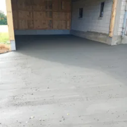 Świeżo wylana, gładka posadzka betonowa wewnątrz drewnianej konstrukcji w stanie surowym, z widocznymi śladami po narzędziach do rozprowadzania betonu, małe okna w ścianie bocznej.