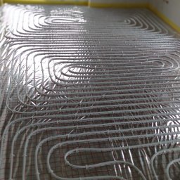 H&C Tomasz Głowacki - Białe rurki systemu ogrzewania podłogowego rozłożone na folii odblaskowej z czerwoną siatką, widok w perspektywie z oknem w tle.