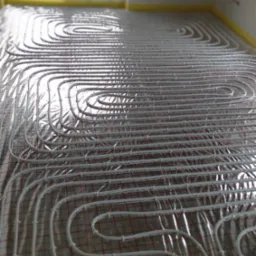 Białe rurki systemu ogrzewania podłogowego rozłożone na folii odblaskowej z czerwoną siatką, widok w perspektywie z oknem w tle.