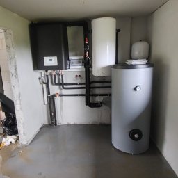 H&C Tomasz Głowacki - Kotłownia w surowym stanie: czarny piec, biały zasobnik ciepłej wody i szary zbiornik buforowy, instalacja rur z zaworami, betonowa podłoga z plamami wody, ściany w stanie deweloperskim, widoczne...