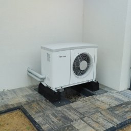 H&C Tomasz Głowacki - Biała pompa ciepła marki Stiebel Eltron zamontowana na czarnych podstawach na szarej kostce brukowej przy białej ścianie budynku.