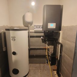 H&C Tomasz Głowacki - Bojler Kospel i szara pompa ciepła w kotłowni z szarymi płytkami, widoczne rury instalacyjne i otwarta przestrzeń za drzwiami.