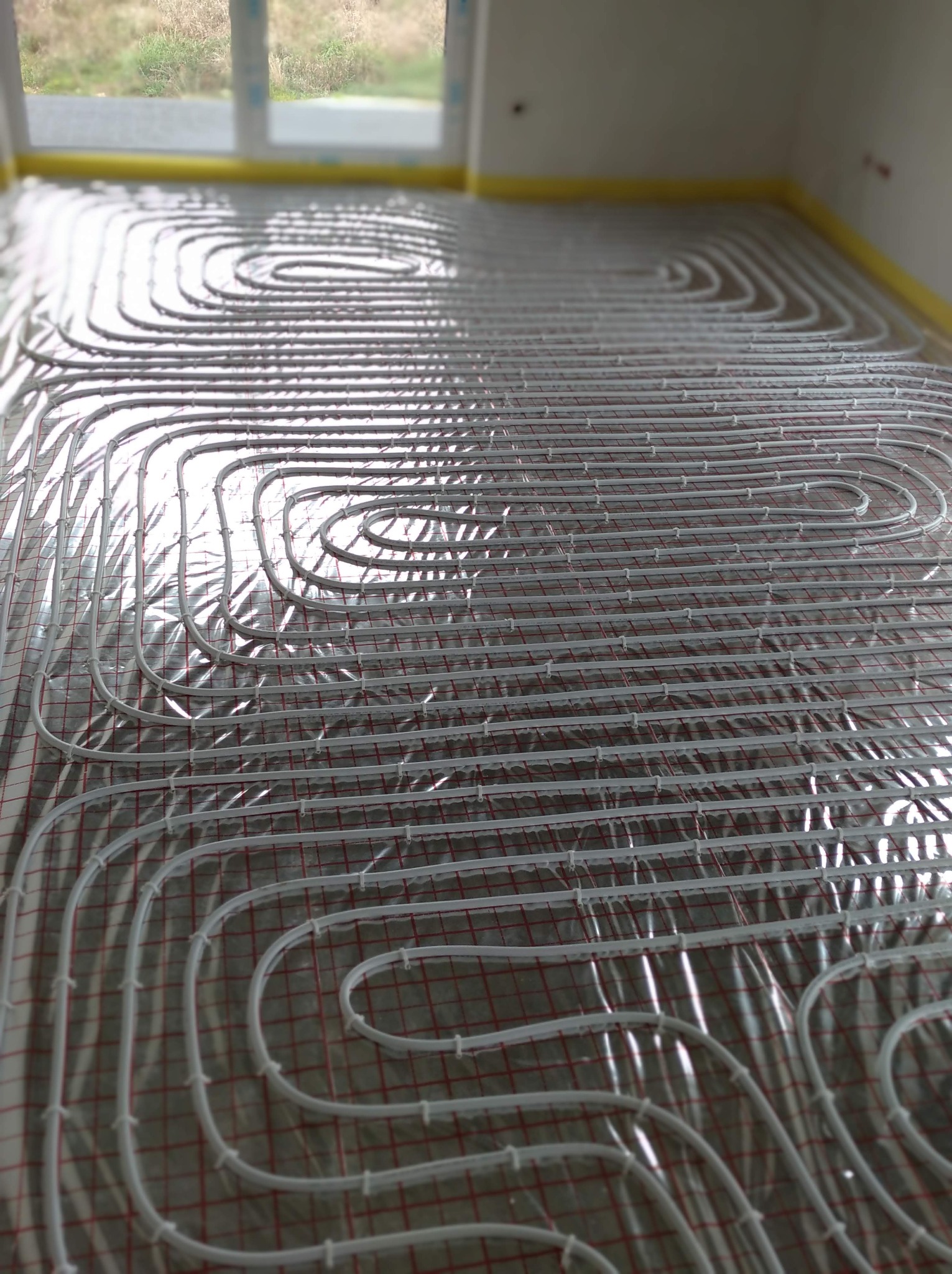Białe rurki systemu ogrzewania podłogowego rozłożone na folii odblaskowej z czerwoną siatką, widok w perspektywie z oknem w tle.