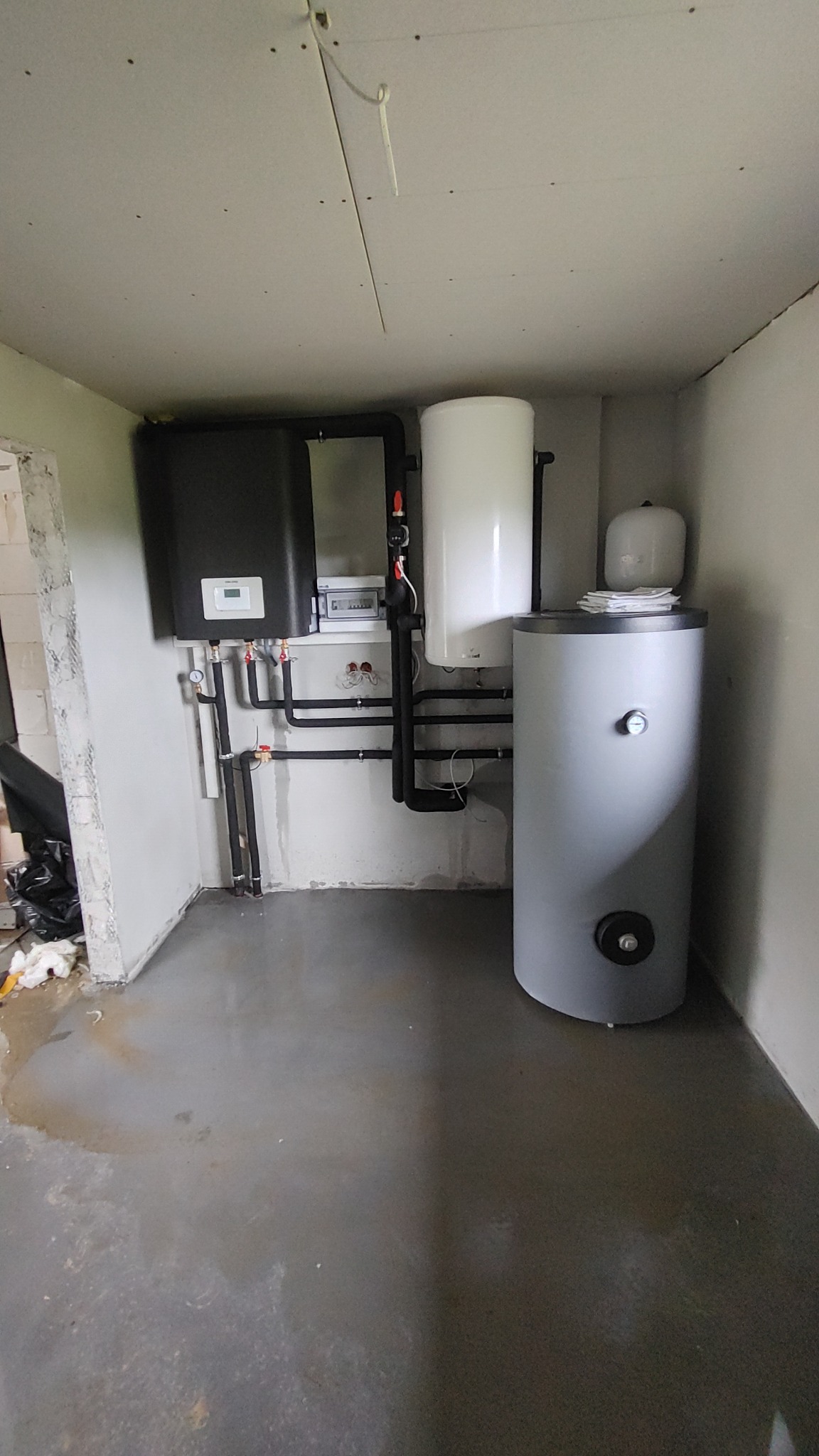 Kotłownia w surowym stanie: czarny piec, biały zasobnik ciepłej wody i szary zbiornik buforowy, instalacja rur z zaworami, betonowa podłoga z plamami wody, ściany w stanie deweloperskim, widoczne...