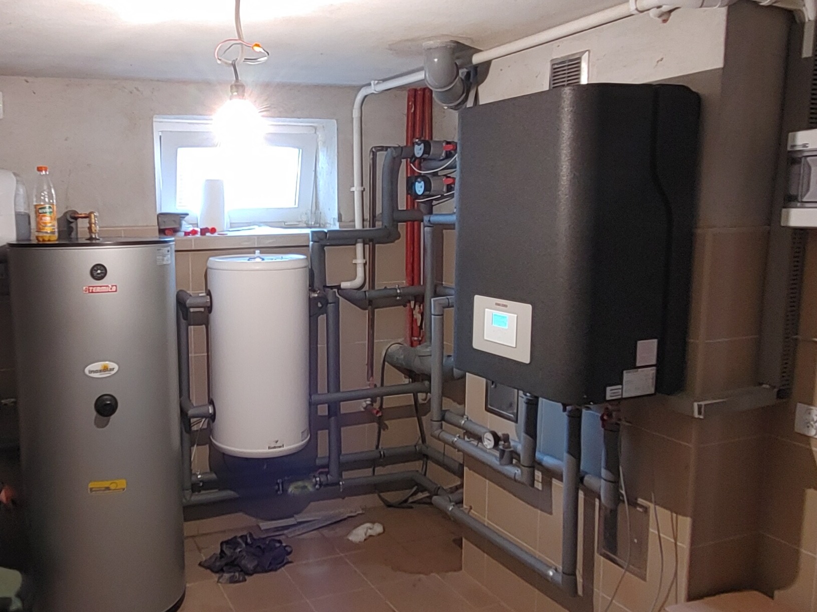 Kotłownia z zainstalowaną pompą ciepła, zbiornikiem buforowym i zasobnikiem ciepłej wody użytkowej. Widoczne liczne rury przyłączeniowe. Na podłodze widoczne ślady po instalacji.