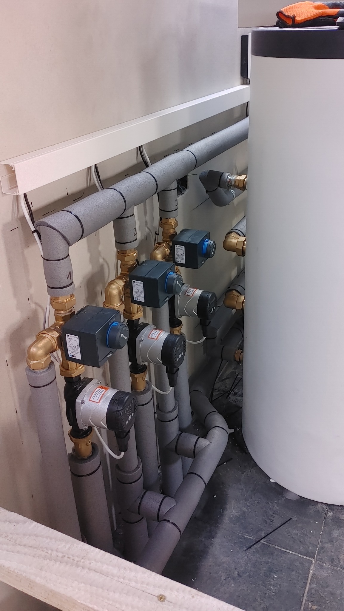 Rozbudowana instalacja z rur w szarej izolacji, zaworów AFRISO i pomp obiegowych zamontowana przy białym zbiorniku akumulacyjnym w pomieszczeniu technicznym.