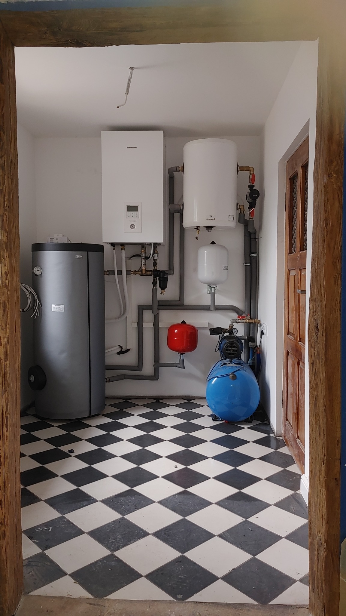 Kotłownia z piecem gazowym, zbiornikiem na wodę, pompą obiegową i naczyniami przeponowymi, widok przez drewniane futryny na czarno-białą podłogę w szachownicę.