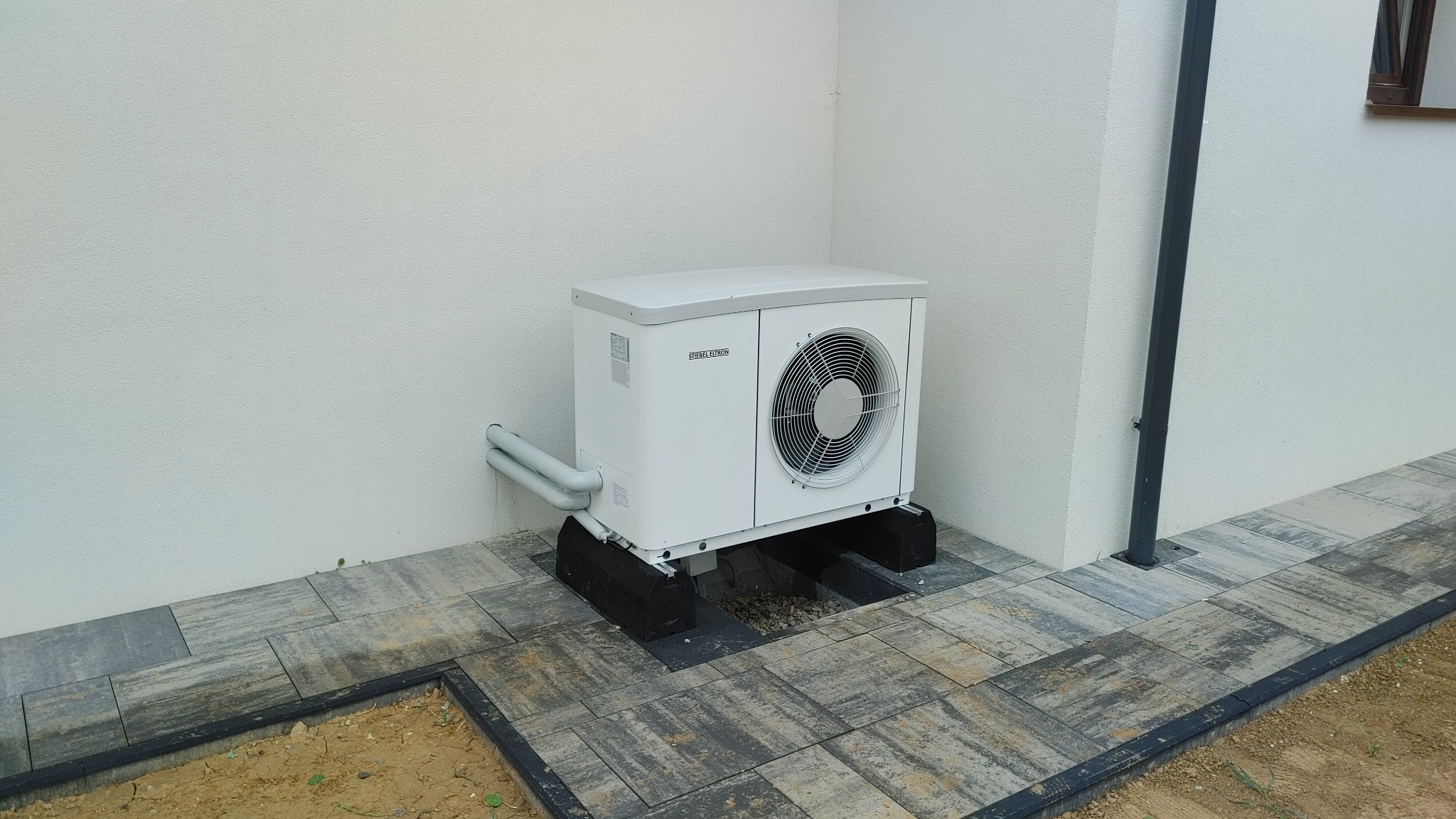Biała pompa ciepła marki Stiebel Eltron zamontowana na czarnych podstawach na szarej kostce brukowej przy białej ścianie budynku.